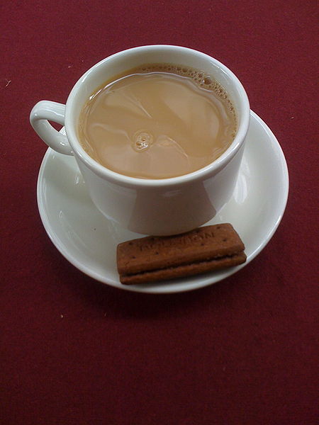 A cup of tea with milk, in a white cup on a white saucer. The saucer also holds two think rectangular cookies. It all sits on a maroon cloth.